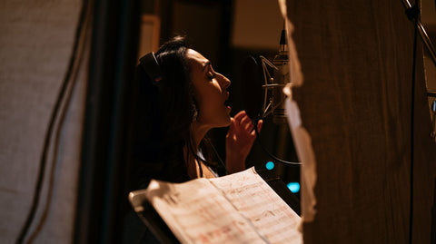 A vocalist sings into a studio microphone while following a marked score in a recording booth.